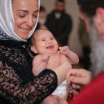 A smiling baby being baptized in a church, held by a woman with a headscarf.